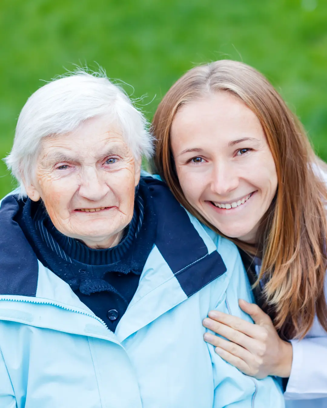 A smiling elderly woman in a light blue jacket poses closely with a young woman, both showing warm expressions amid a green background.