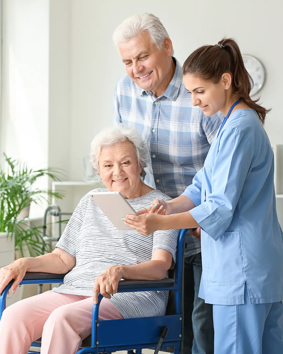 A healthcare professional showing a tablet to an elderly woman seated in a wheelchair, accompanied by a smiling man. The setting is bright and airy, with plants visible in the background.